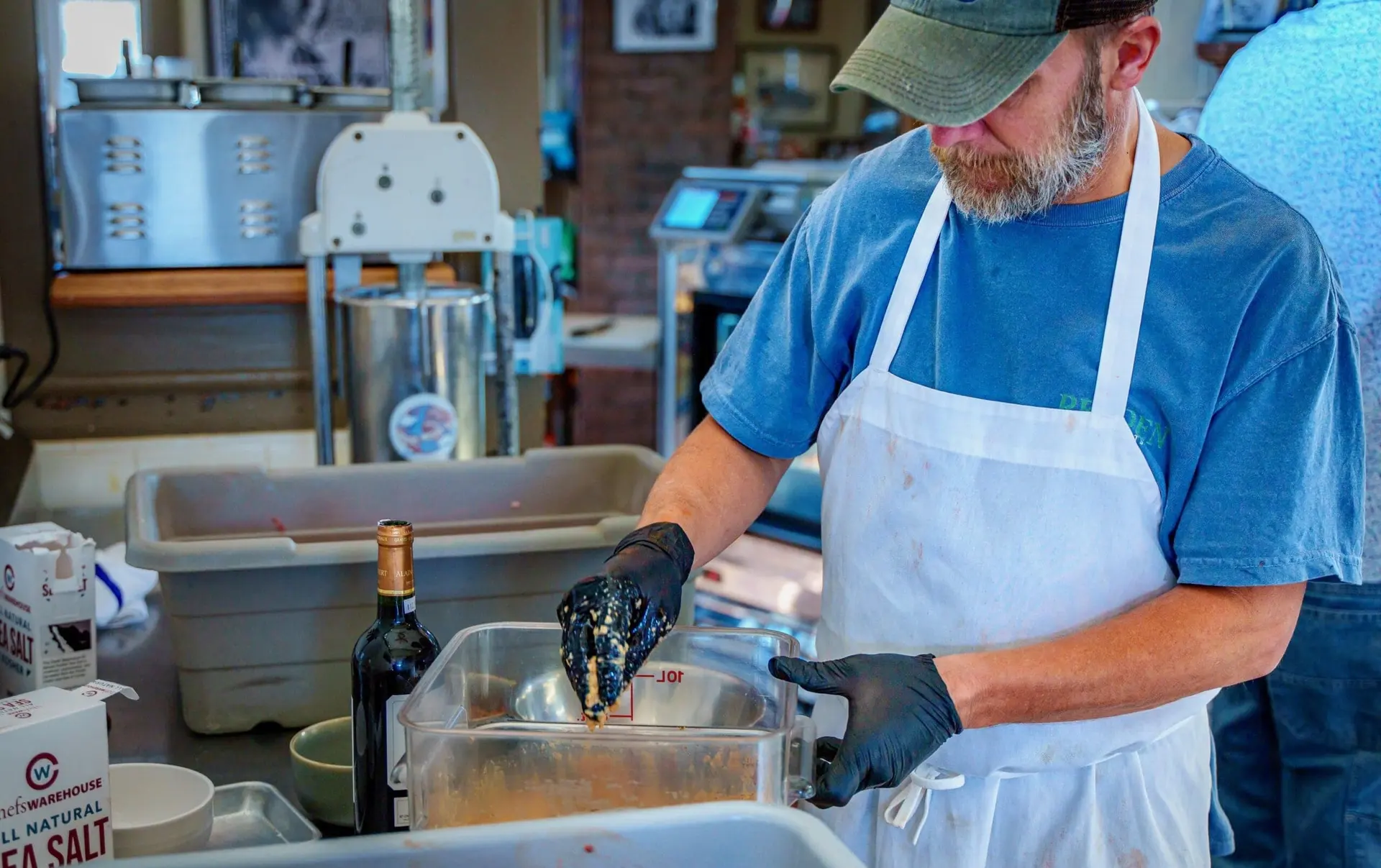 A butcher in a cap, blue T-shirt and white apron seasons a tub of ground meat in the shop’s back kitchen, with sausage-making equipment behind him