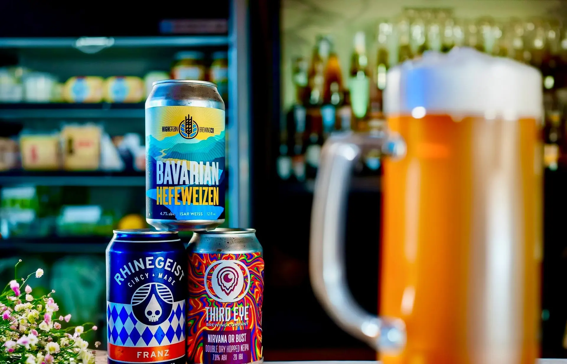 Cans of craft beer stacked beside a frosty mug of beer, with the shop’s shelves blurred behind