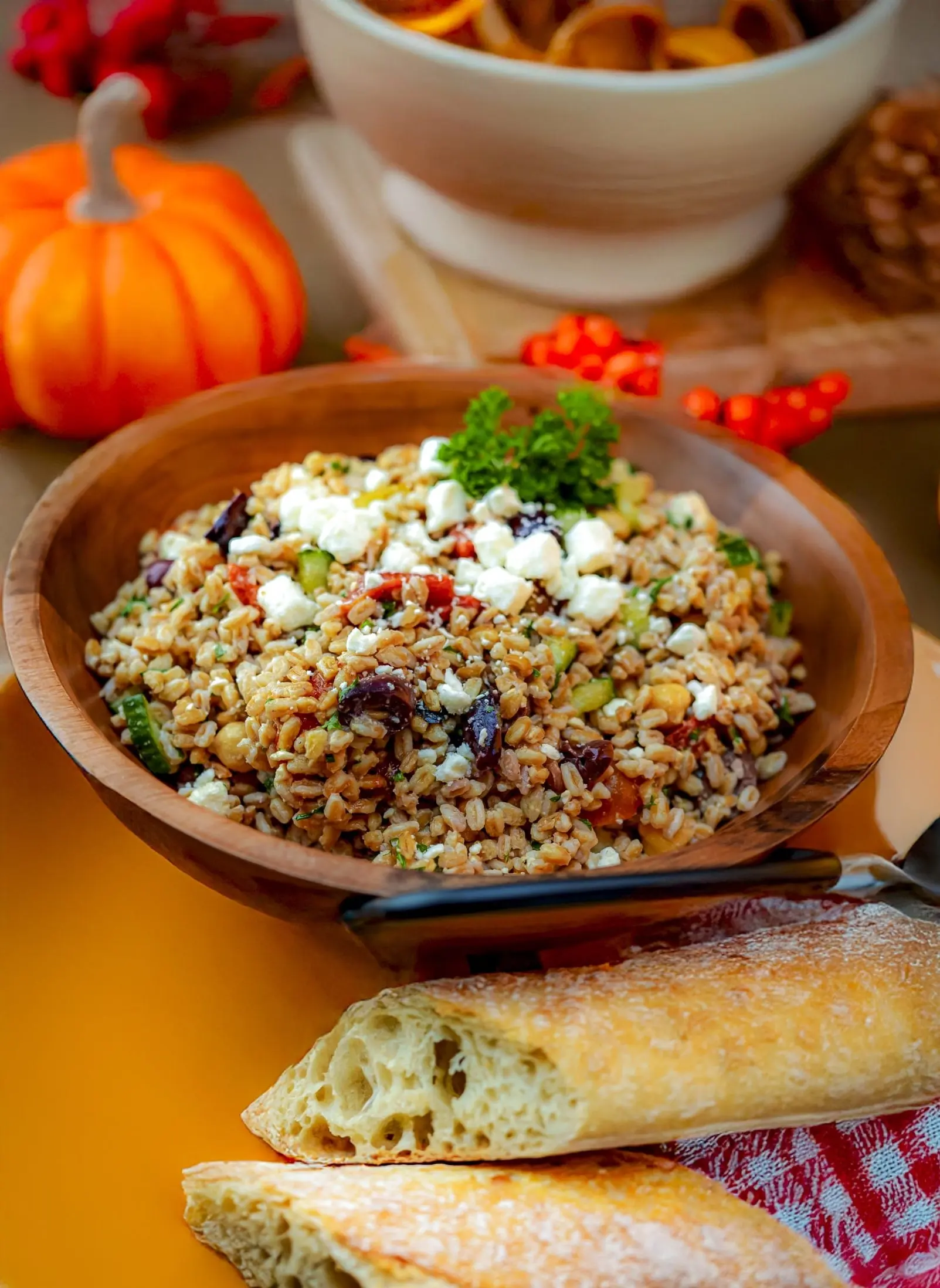 A wooden bowl of farro salad with feta and parsley, with sliced baguette and a small pumpkin on the table
