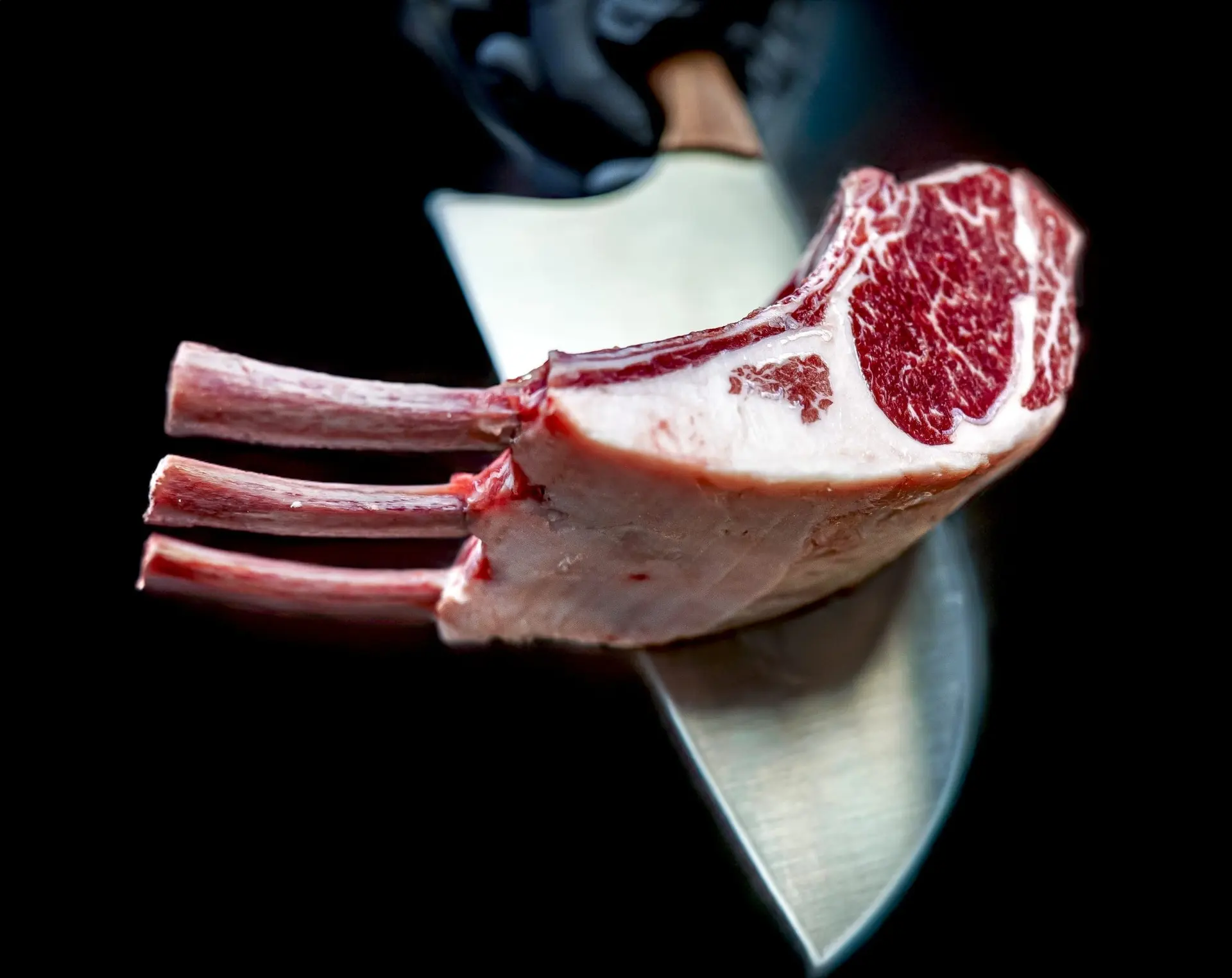 A frenched rack of three raw, deeply marbled chops balanced on the blade of a chef’s knife against a black background