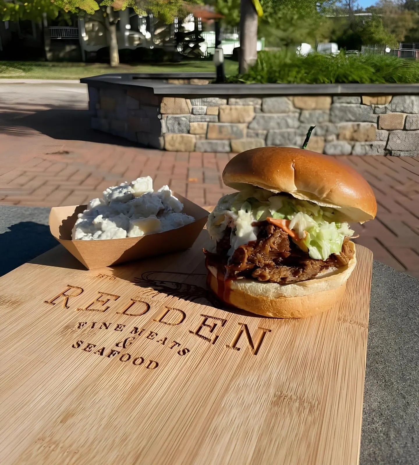 A pulled-meat sandwich with slaw and a deli side on a laser-engraved REDDEN Fine Meats & Seafood cutting board, outdoors on a brick plaza