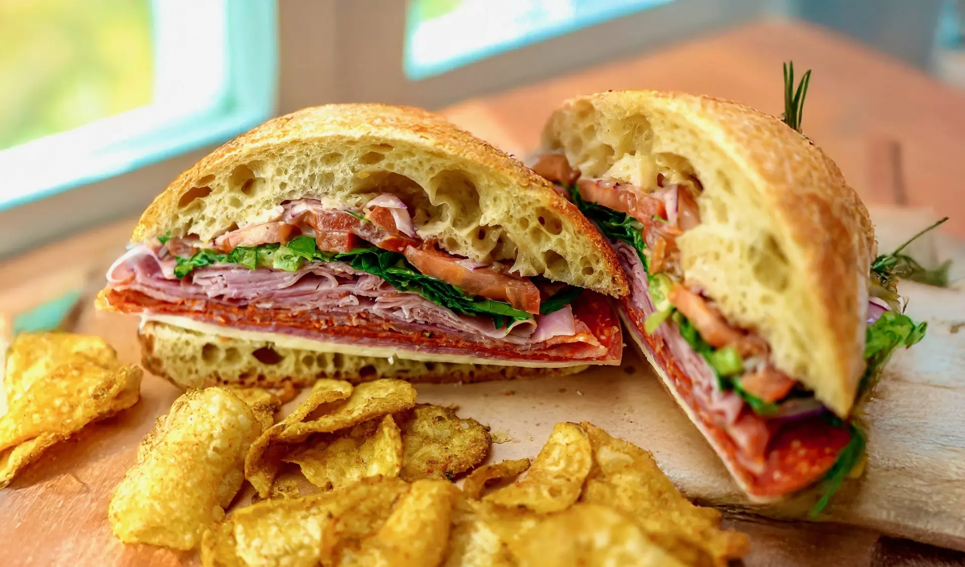 A ciabatta sandwich layered with Italian cured meats, greens and tomato, cut in half beside kettle chips in soft window light