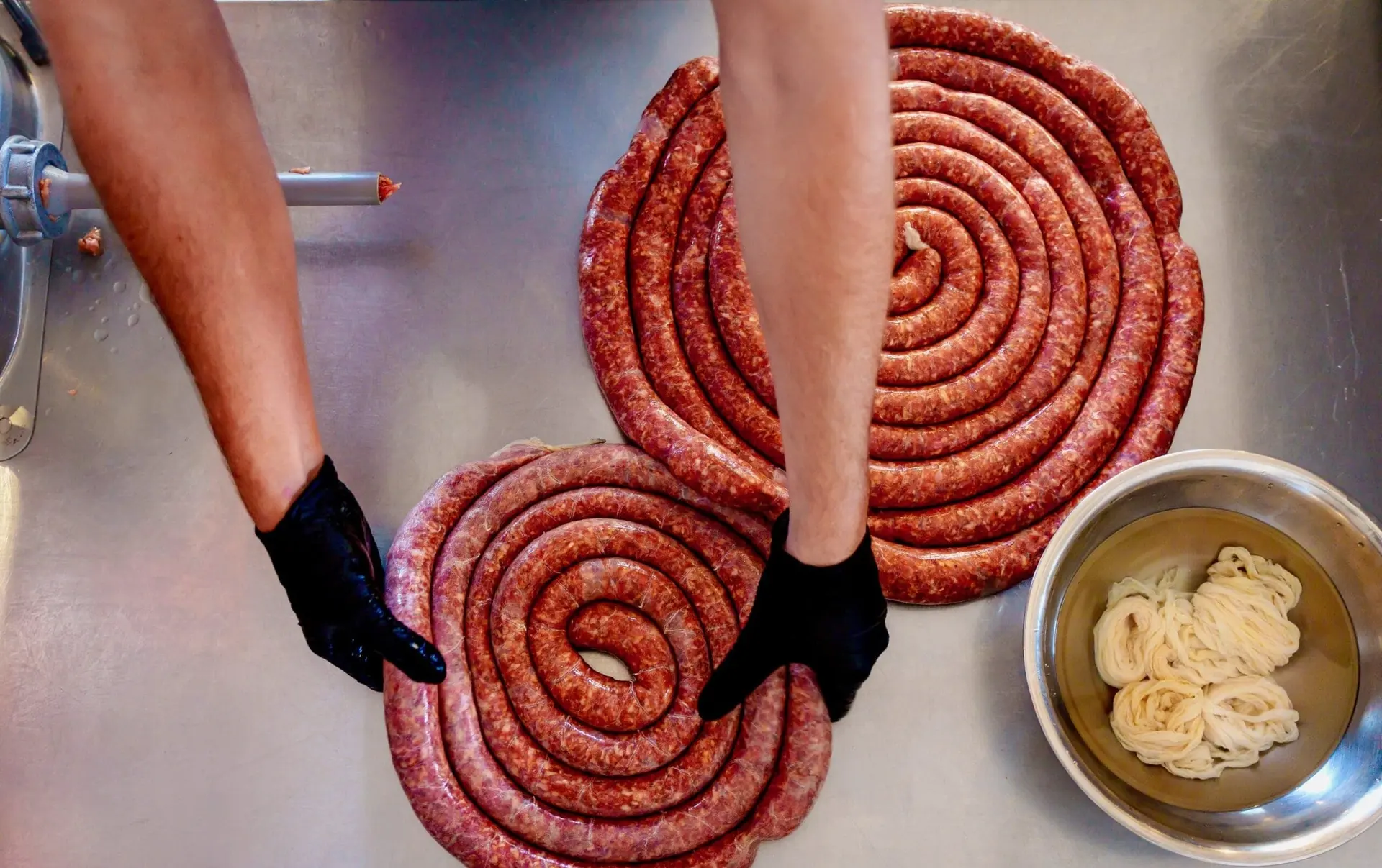 Gloved hands coiling long ropes of fresh sausage into spirals on a steel prep table, beside a bowl of casings