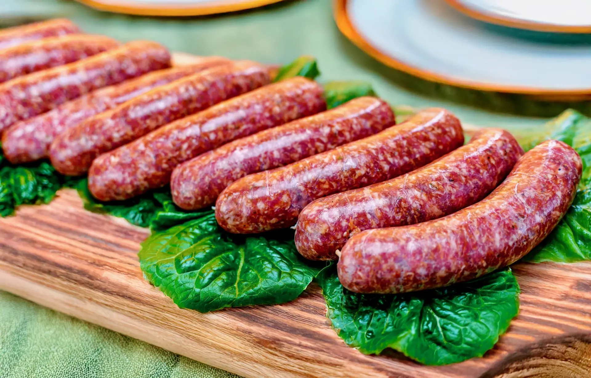 A row of plump raw sausage links on lettuce leaves on an olive-wood board