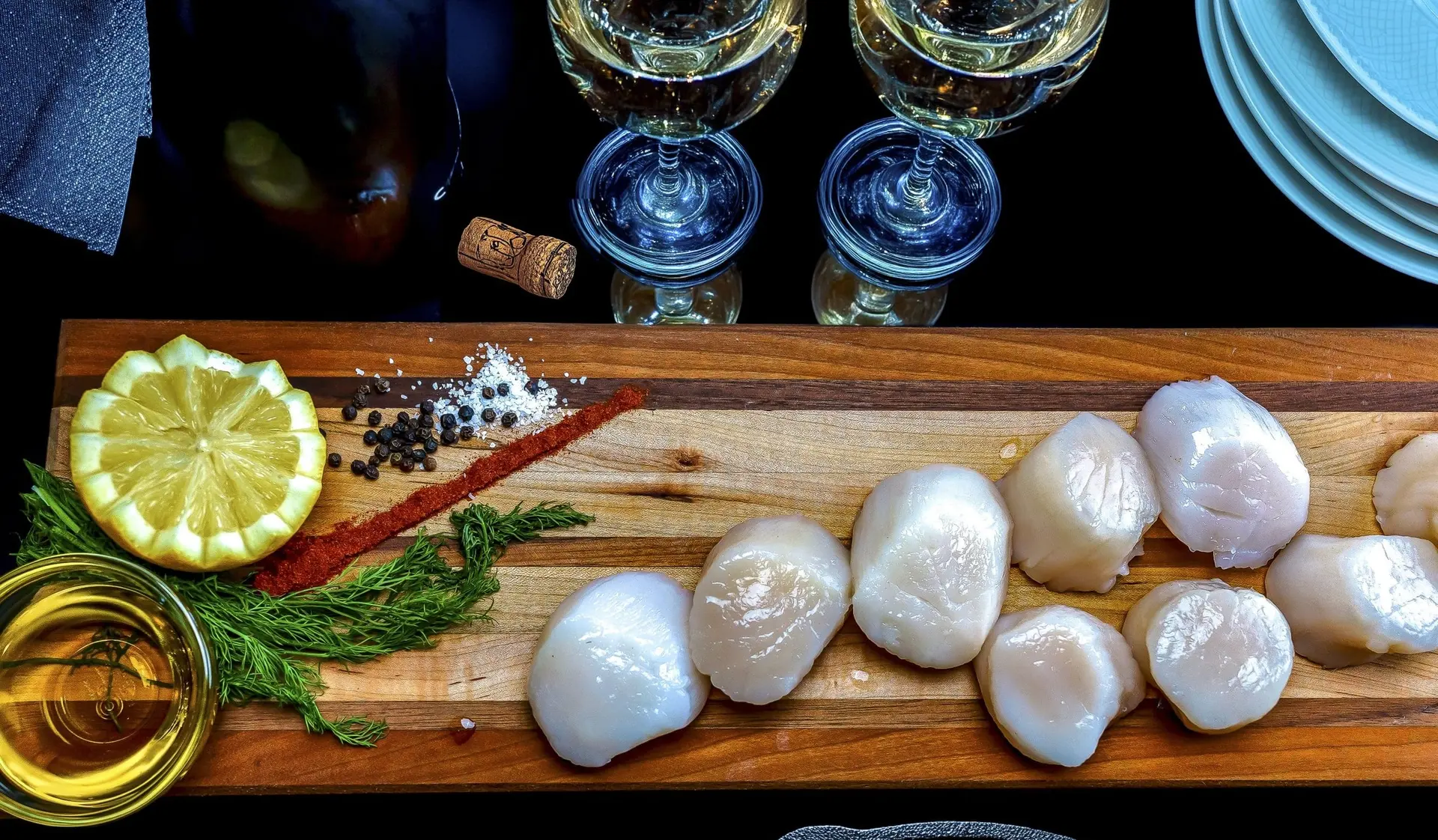 Large raw sea scallops lined up on a narrow wooden board with lemon, dill and peppercorns, wine glasses behind
