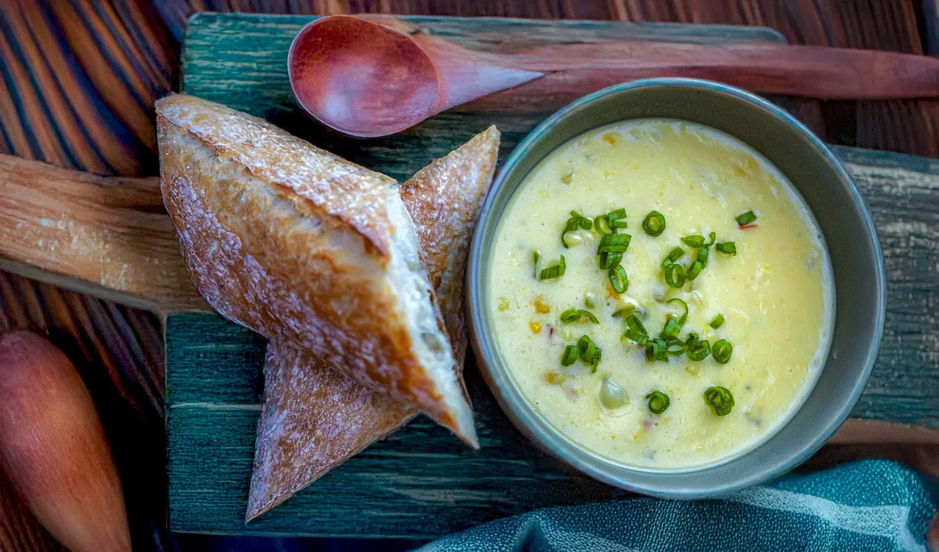 A bowl of creamy soup topped with chives beside a torn baguette on a teal wooden board