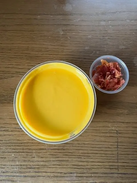 A to-go container of golden soup with a small cup of crispy topping, seen from above on a wooden table